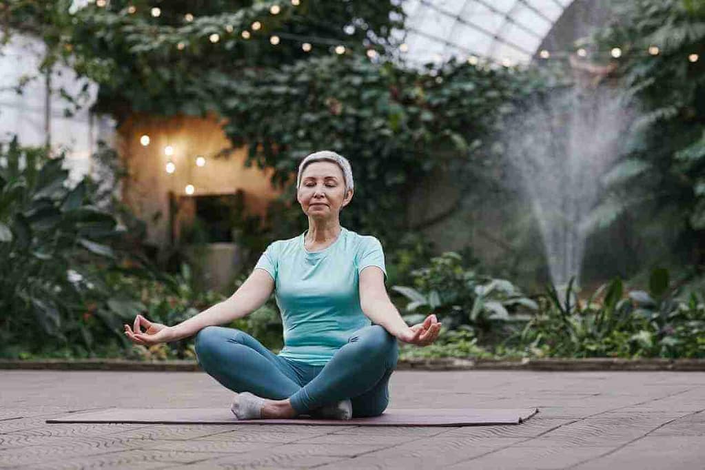 older woman white hair. wearing a blue top and dark pants. sitting crossed legged outside on a yoga mat. she is a beginner at yin yoga.