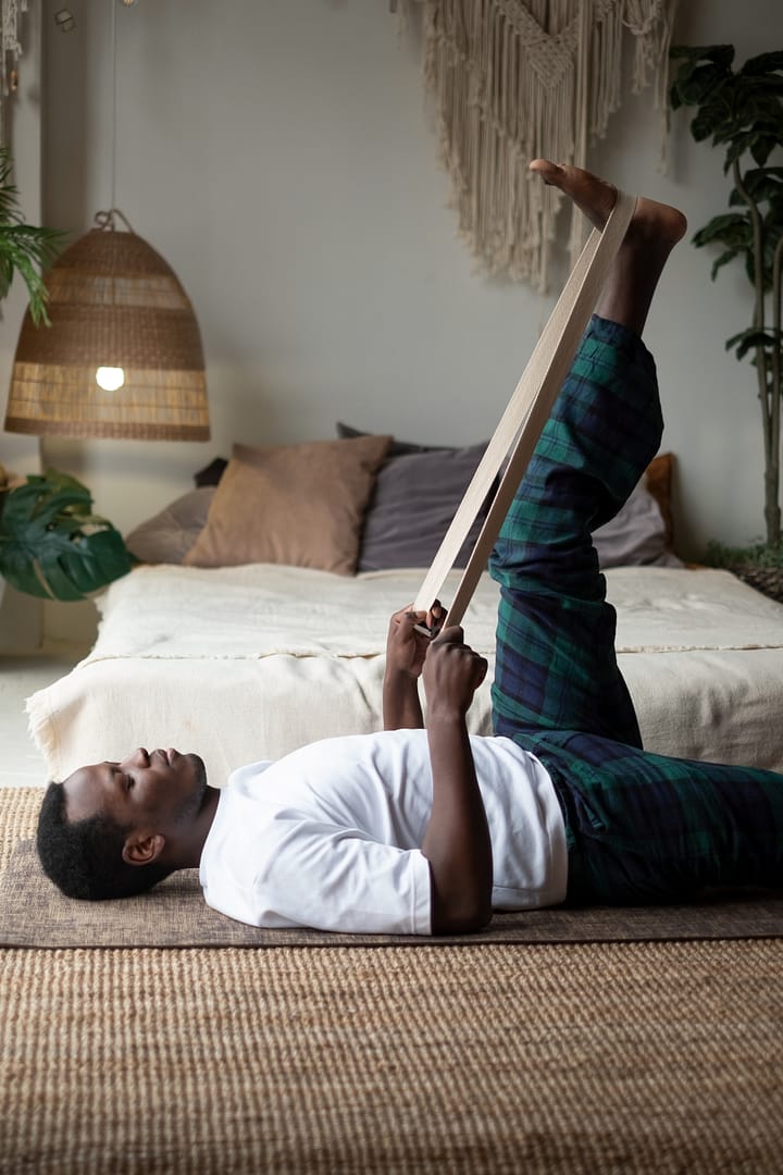 The Benefits of Using Yoga Props: Young african man working out at home , warming up using yogic belt, lying in yoga Supta Padangushthasana, One Leg Lift exercise, reclined variation of Extended Hand to Big Toe pose