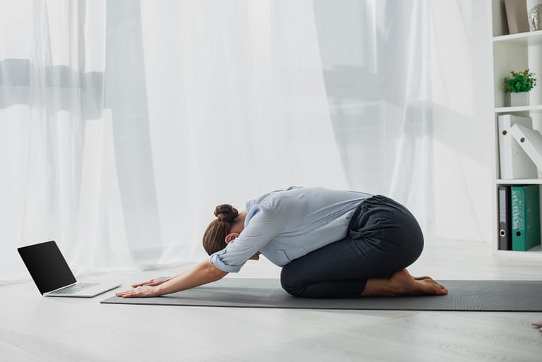woman at home doing a 30 day yoga challenge performing child pose in casual clothing in front of computer. childs pose is a counterpose to anahatasana pose melting heart pose yin yoga