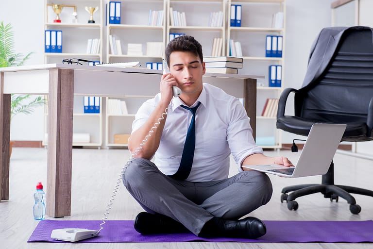 man sitting on a yoga mat on the floor of an office holding a phone. taking advantage of benefits of yoga in the workplace