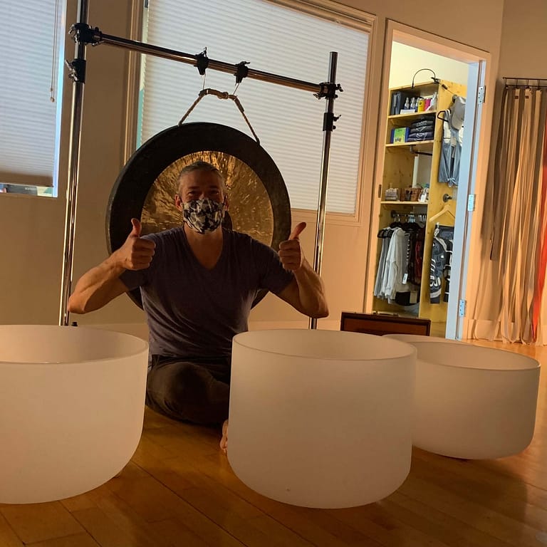 man with mask on sitting in behind crystal bowls with a gong in the back ground. hosting a sound bath meditation